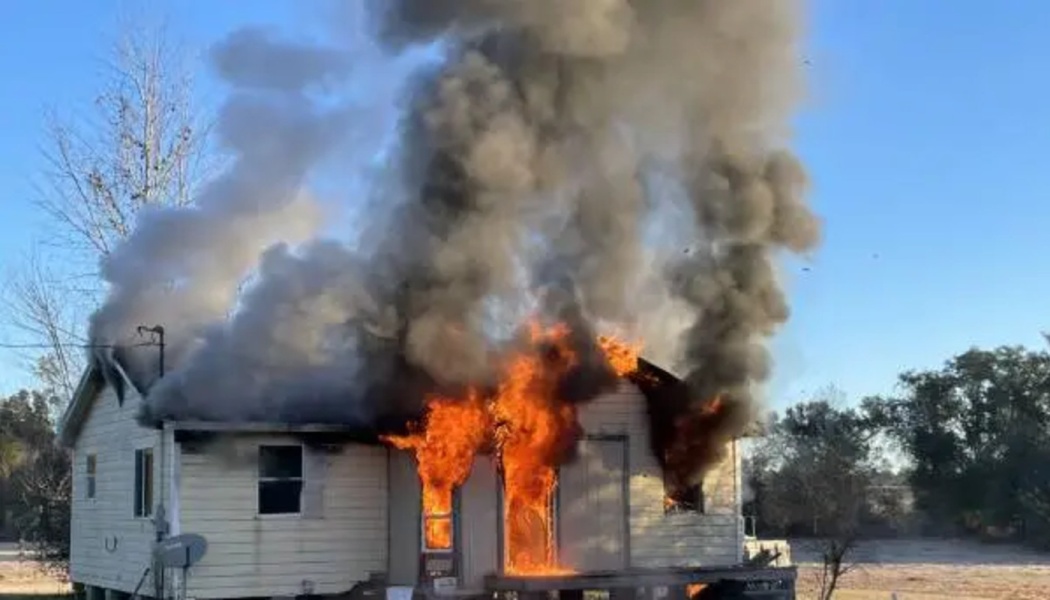 A single-story house engulfed in bright orange flames and heavy dark smoke.