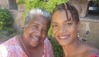 Two smiling Black women pose warmly together outdoors in front of lush green plants.