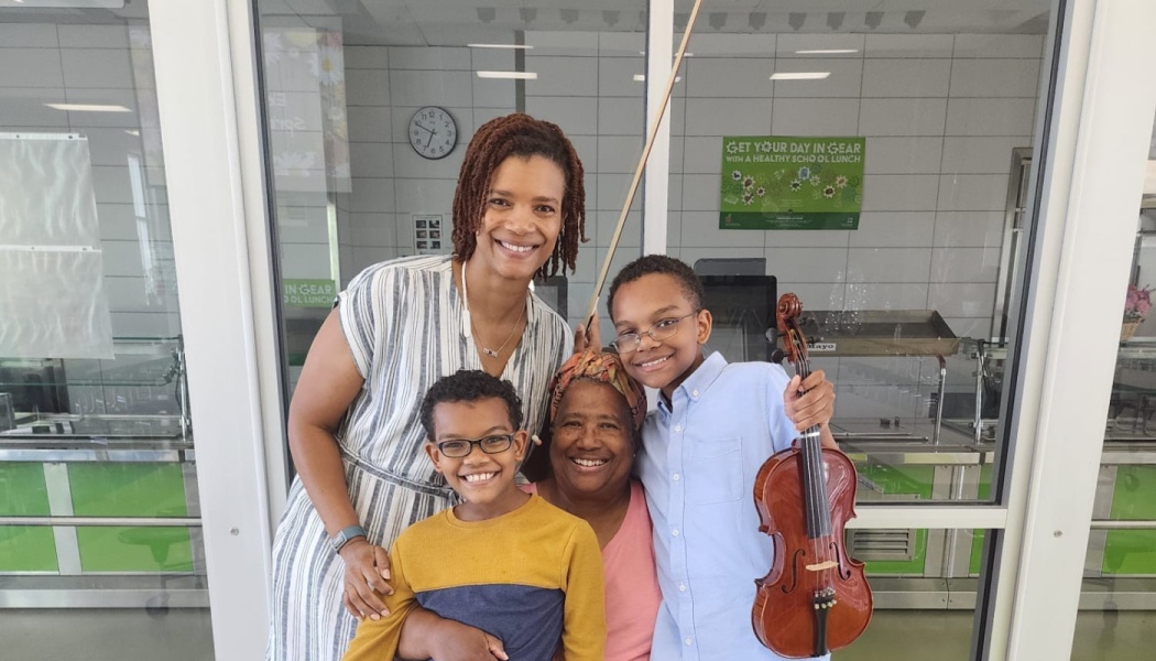 A smiling mother, grandmother, and two young boys pose together, with one boy holding a violin and bow.