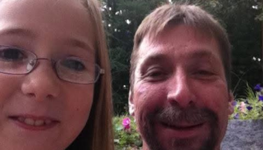 A young girl with glasses and a smiling man with a goatee posing for a close-up selfie outdoors.