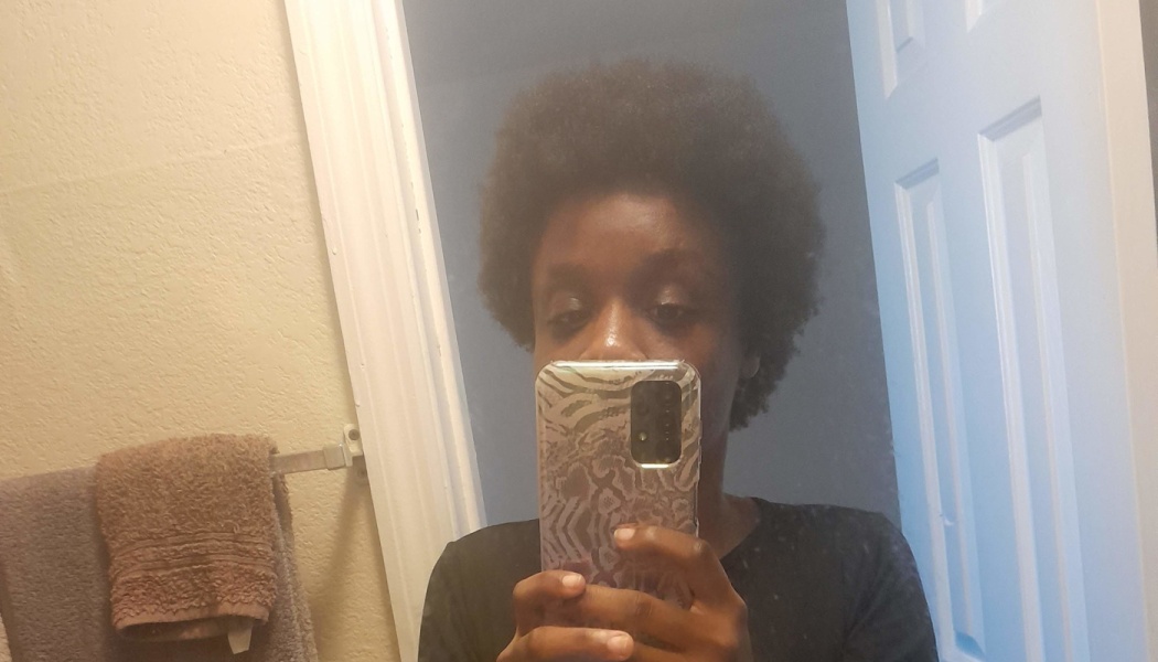 Person with short afro hair holding a patterned smartphone to take a mirror selfie in a bathroom.