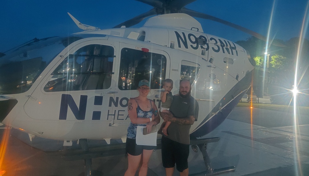 A family with a toddler posing together in front of a Novant Health medical helicopter at night.