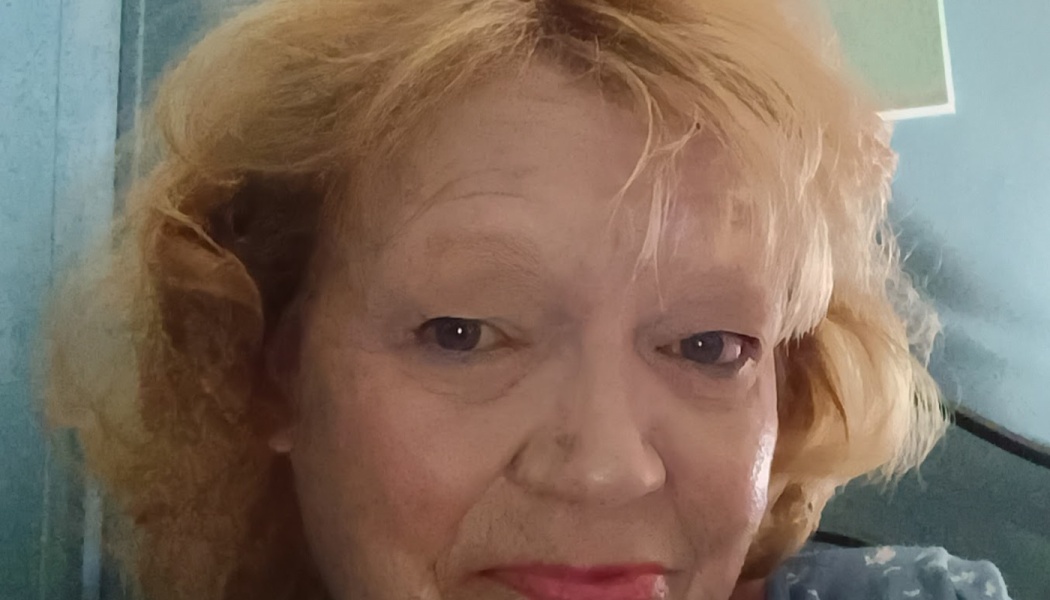 Close-up portrait of an older woman with short blonde hair and red lipstick looking into the camera.