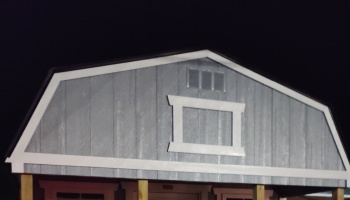 Upper front facade of a grey wooden shed featuring white trim, a decorative center frame, and gambrel roofline.