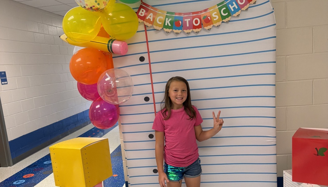 A smiling girl poses with a peace sign in front of a Back to School notebook backdrop with colorful balloons.