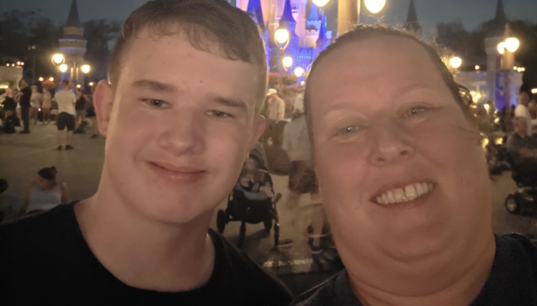 A smiling woman and teenage boy take a selfie at night in front of an illuminated Disney castle.