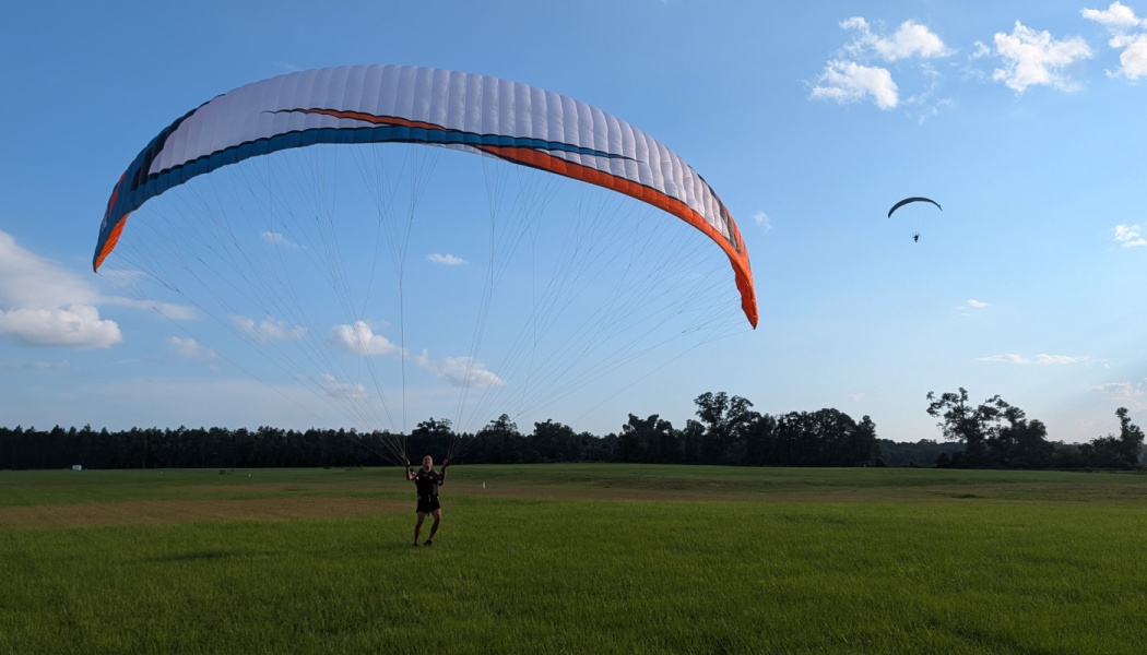A paramotor pilot inflates a white and orange paraglider wing in a grassy field under a blue sky.