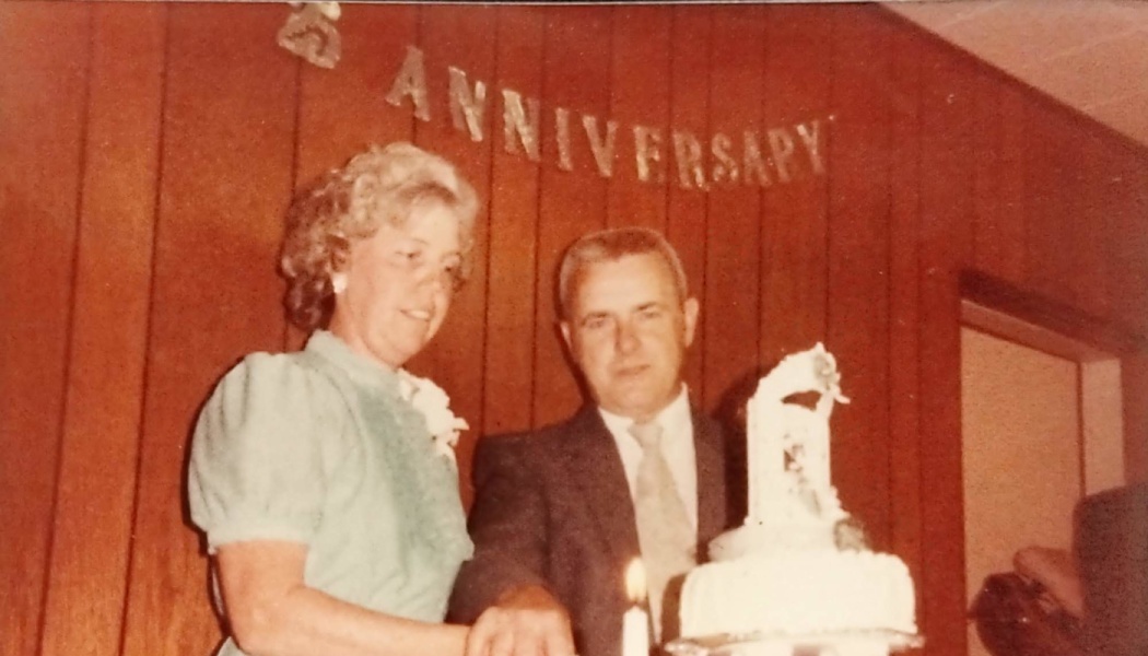 An older couple stands together cutting a tiered cake under a 25th Anniversary banner.