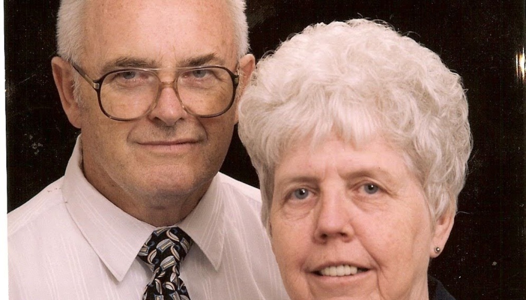 An elderly man with glasses and a tie poses closely with an elderly woman with short white hair.