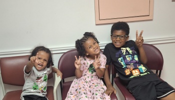 Three happy children sitting together in chairs, making peace signs and playful poses for the camera.