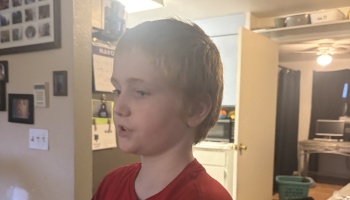 Young boy with short brown hair wearing a red shirt looking slightly to the side indoors.