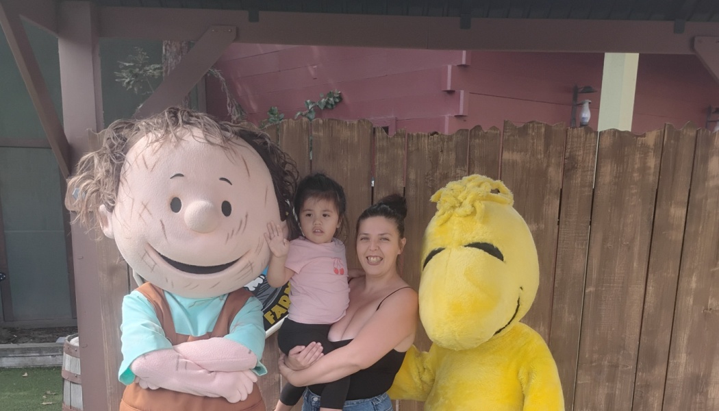 A woman holding a young child poses with Peanuts costumed characters Pig-Pen and Woodstock.