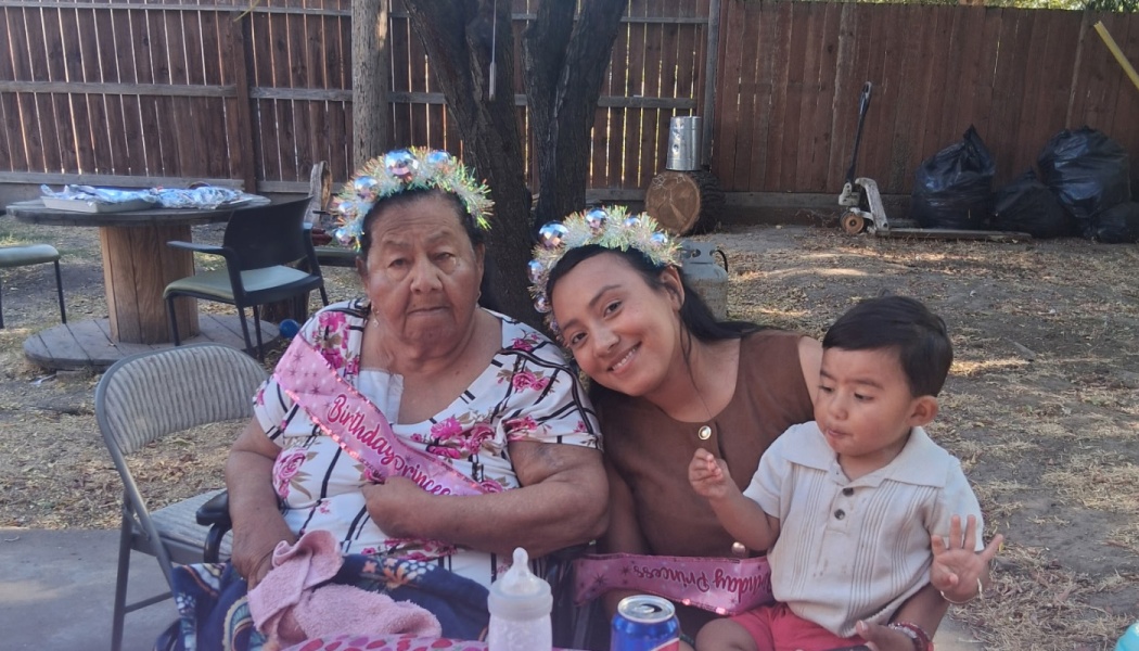 An elderly woman wearing a birthday sash and tiara sits outdoors with a smiling younger woman and a toddler.