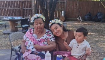 An elderly woman wearing a birthday sash and tiara sits outdoors with a smiling younger woman and a toddler.