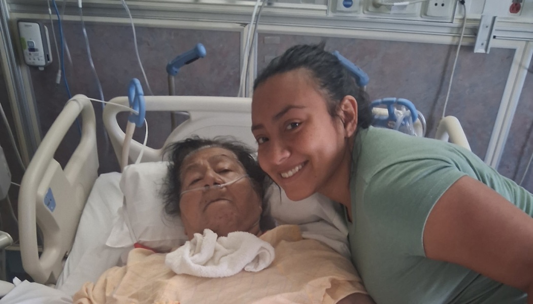 A smiling young woman poses next to an elderly woman lying in a hospital bed with a nasal cannula.