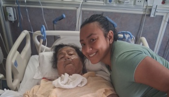 A smiling young woman poses next to an elderly woman lying in a hospital bed with a nasal cannula.