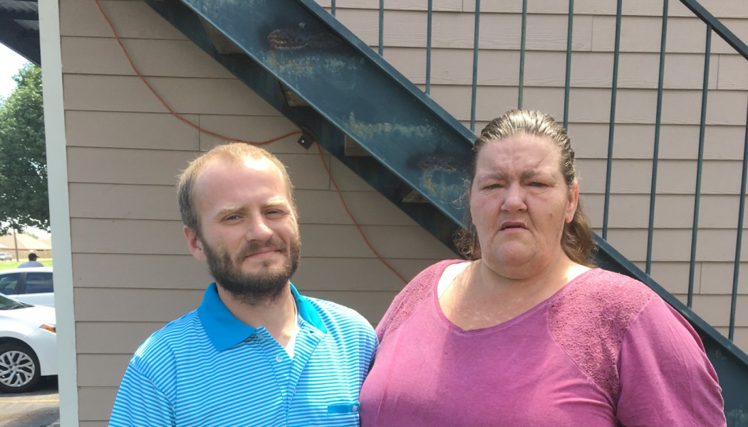 A bearded man in a blue striped shirt and a woman in a pink top standing together outside an apartment building.