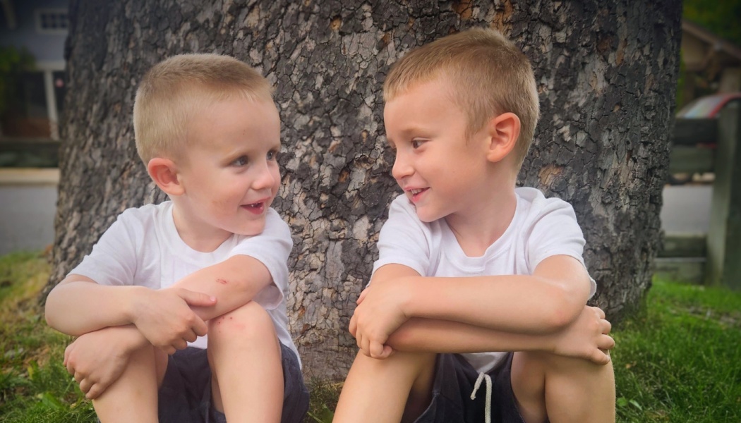 Two young boys in matching white t-shirts sit against a tree trunk, looking at each other and smiling.