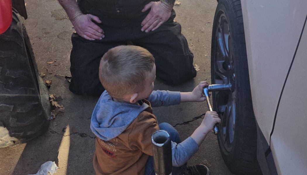 A young boy uses a wrench on a car tire while an adult kneels nearby supervising.