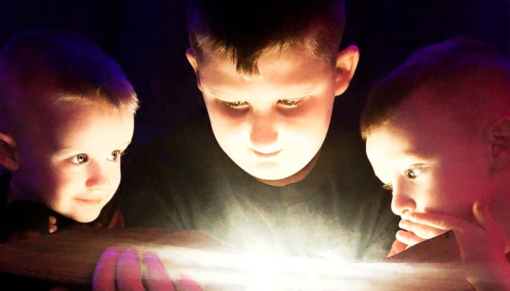 Three young boys looking down in wonder at a bright light illuminating their faces in the dark.
