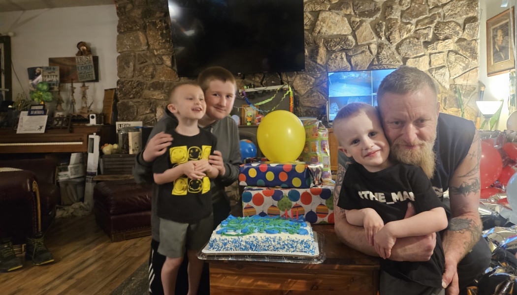 A smiling father and three young boys gather around a blue birthday cake, wrapped presents, and a yellow balloon.