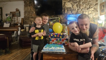 A smiling father and three young boys gather around a blue birthday cake, wrapped presents, and a yellow balloon.