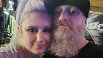 Close-up selfie of a blonde woman leaning close to a man with a long beard and black cap.