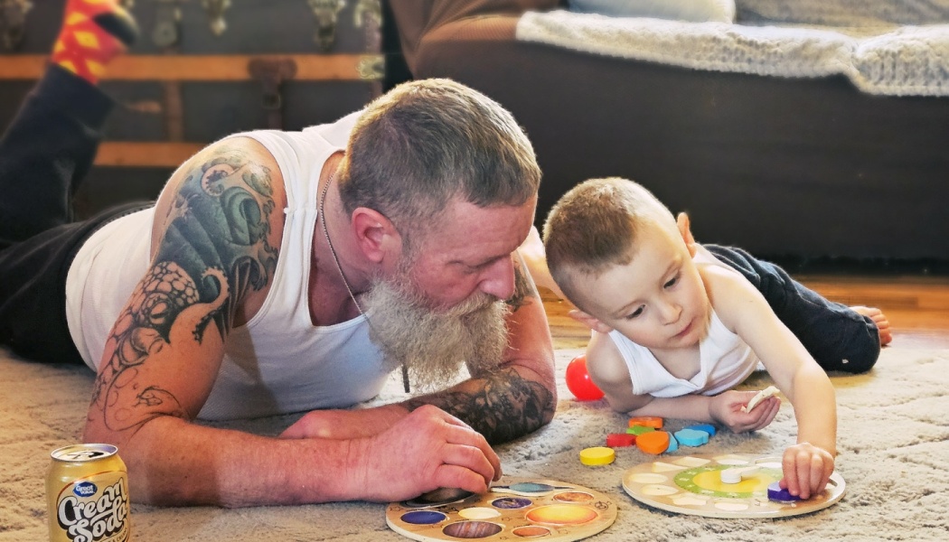 A tattooed man with a gray beard lies on a rug playing with wooden puzzles alongside a young boy.