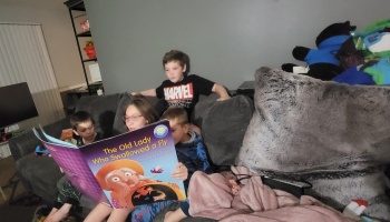 A young girl reads a large storybook titled "The Old Lady Who Swallowed a Fly" to three children on a couch.