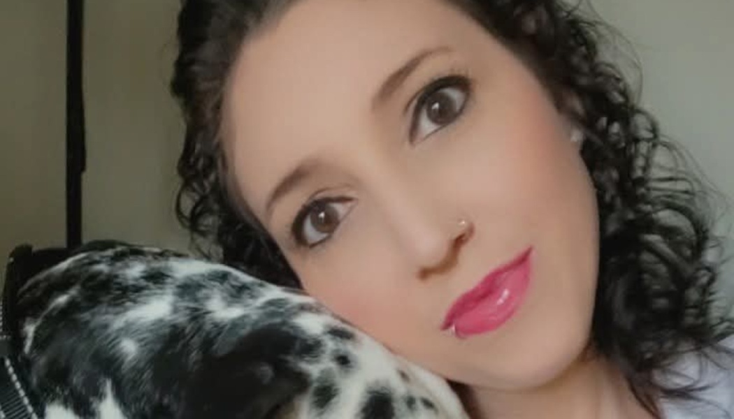 A woman with dark curly hair and pink lipstick resting her head on a black and white spotted dog.