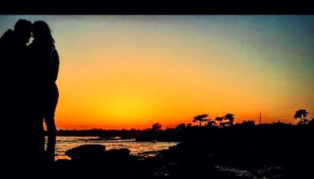 Silhouette of a couple standing close together against a vibrant orange sunset over the ocean.