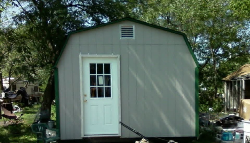 Small light gray barn-style storage shed with dark green trim and a white front door in a wooded yard.