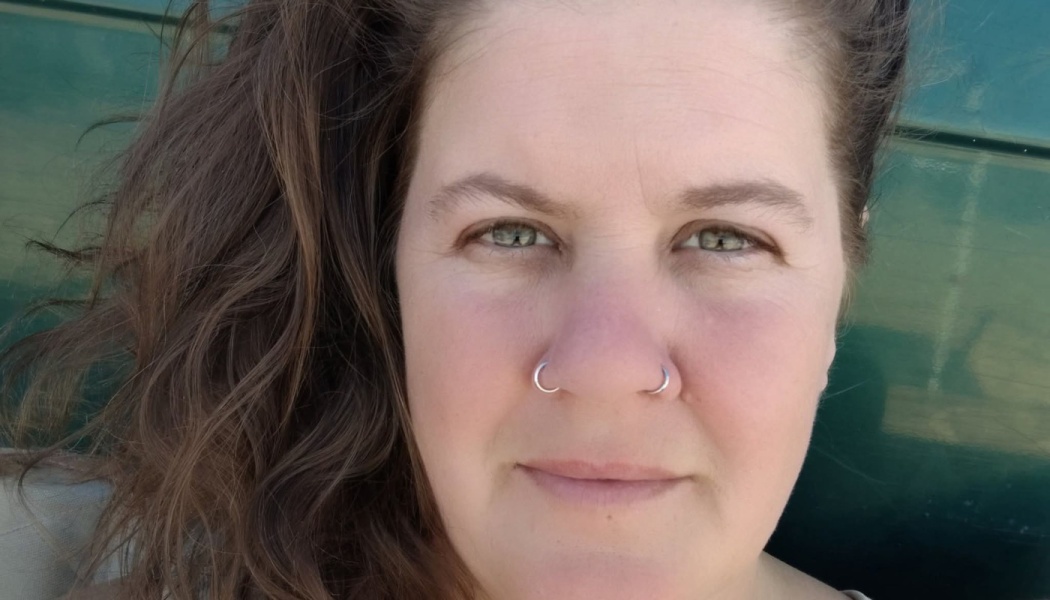 Close-up portrait of a woman with wavy brown hair, hazel eyes, and silver nostril rings.