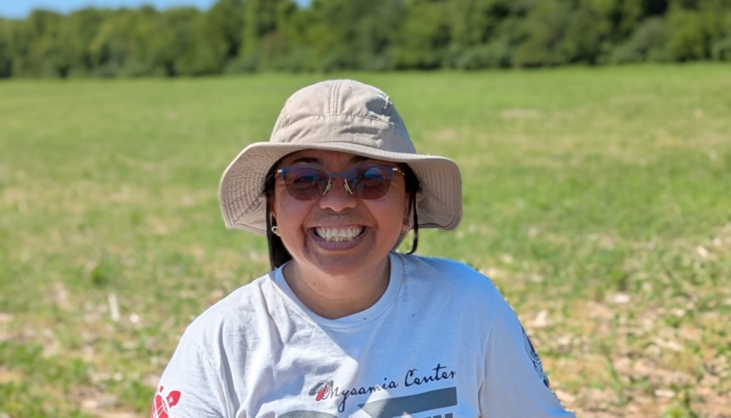 A smiling woman wearing a sun hat, sunglasses, and white t-shirt outdoors in a grassy field.