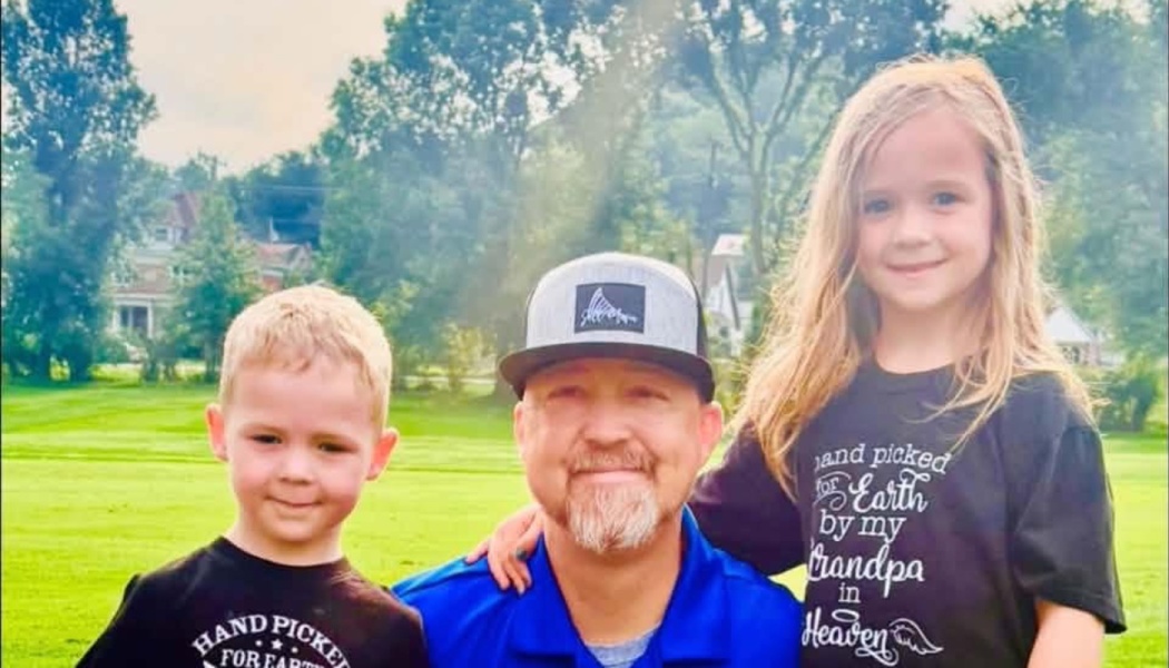 A smiling man in a baseball cap poses outdoors with a young boy and girl in a grassy park.