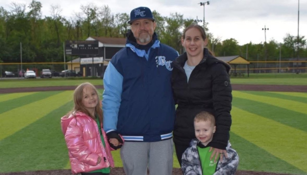 A smiling family of four standing together on a baseball field.