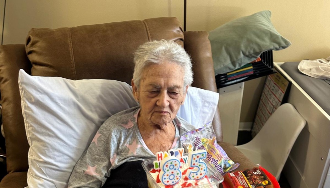 An elderly woman resting in a brown recliner, looking at a small cake with 85th birthday candles on her lap.