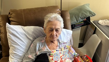 An elderly woman resting in a brown recliner, looking at a small cake with 85th birthday candles on her lap.