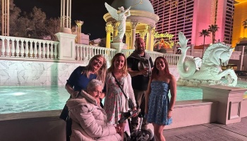 A smiling family, including an elderly woman in a wheelchair, poses in front of a lit Las Vegas fountain at night.