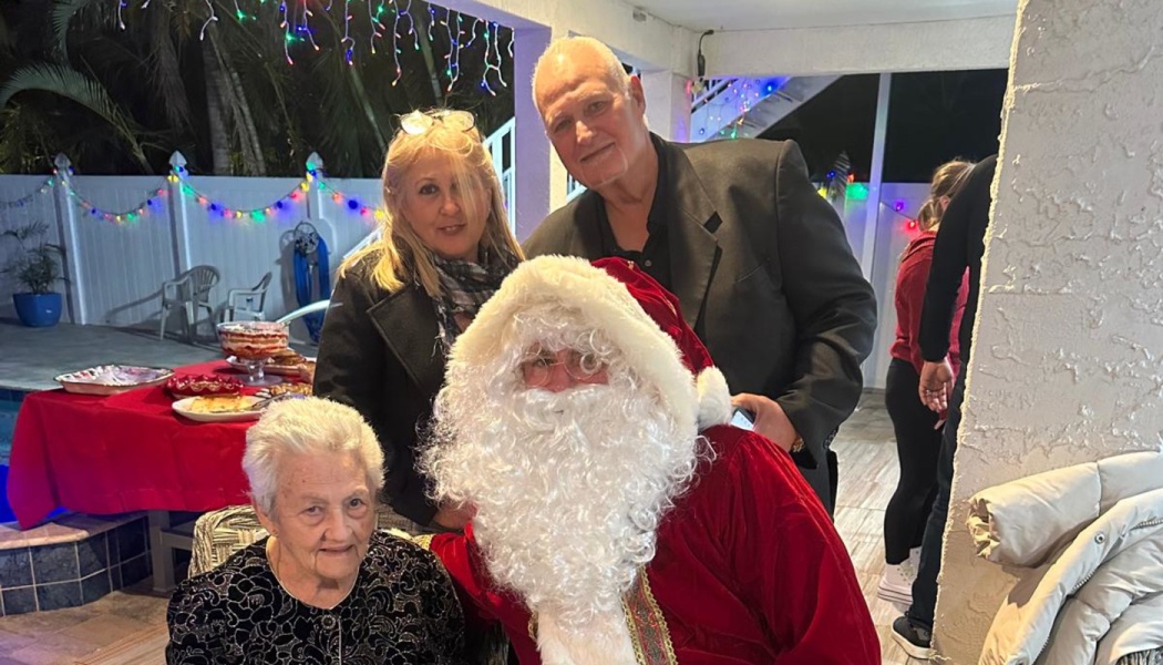 An elderly woman sitting beside Santa Claus with a couple standing behind them at a festive holiday party.