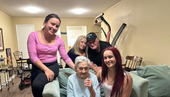 A smiling multi-generational family gathered around a senior woman sitting on a couch at home.