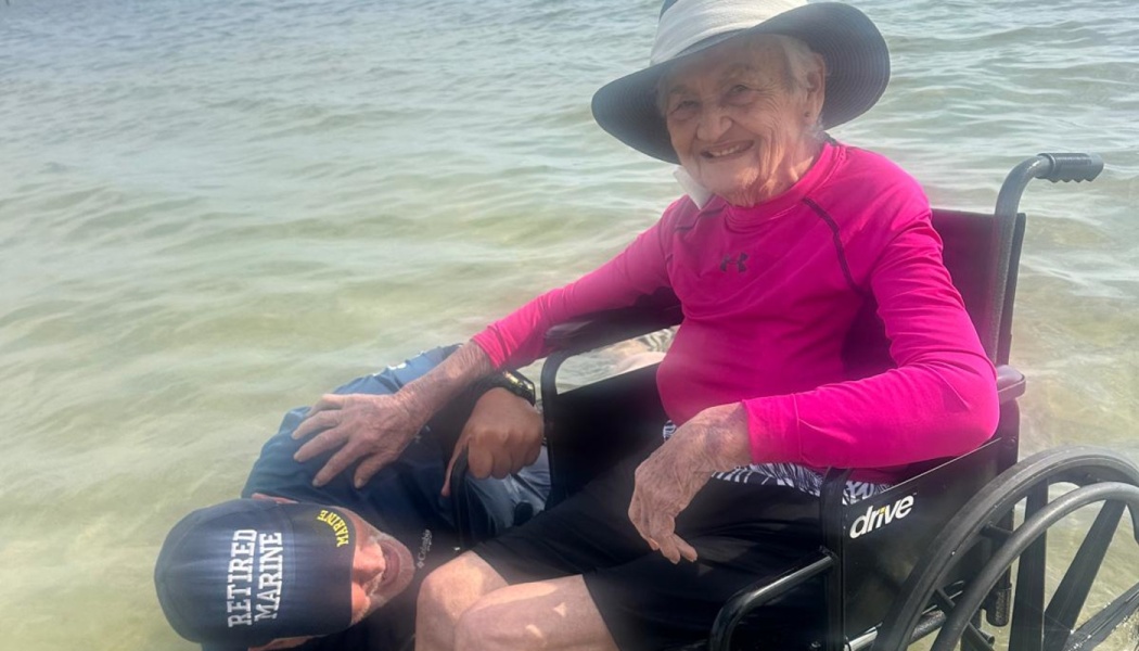 Smiling elderly woman in a wheelchair enjoys shallow ocean water next to a man wearing a Retired Marine cap.
