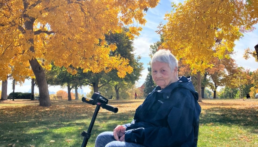 An elderly woman sits on a mobility scooter in a park surrounded by vibrant golden autumn trees.