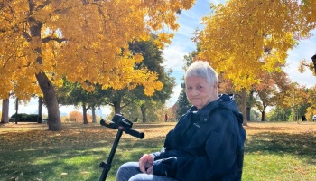 An elderly woman sits on a mobility scooter in a park surrounded by vibrant golden autumn trees.
