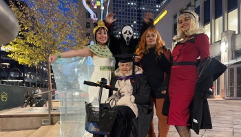 A group of people in Halloween costumes pose at night around an elderly woman sitting in a mobility scooter.