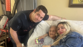 A man and a blonde woman pose affectionately with an elderly woman resting in a bed.
