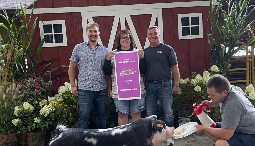 Three people pose with a Breed Champion award banner behind a black and white spotted pig at a county fair.