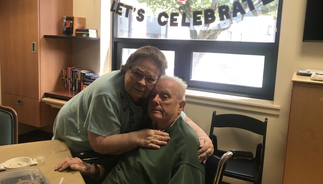 An elderly woman tenderly hugs a senior man sitting in a wheelchair under a 'LET'S CELEBRATE' banner.
