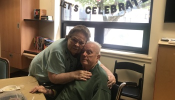 An elderly woman tenderly hugs a senior man sitting in a wheelchair under a 'LET'S CELEBRATE' banner.