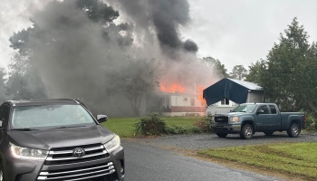 Flames and heavy dark smoke engulf a residential home with vehicles parked in front.